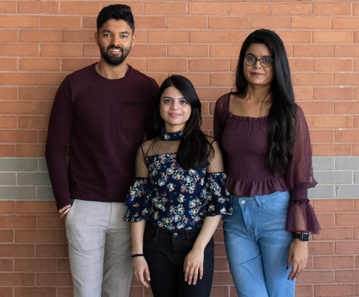 Aakash, Bhumi, and Shreya stand together against a red and grey brick background on UArizona Campus