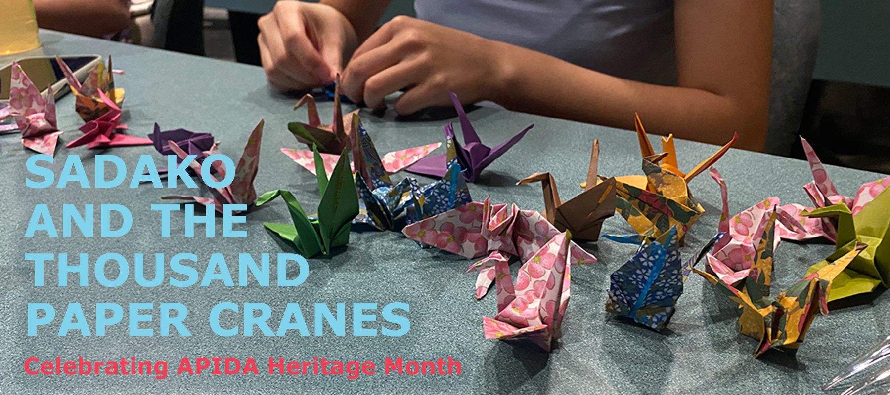Person's hands are shown making origami paper cranes with 20 colorful cranes on table already constructed