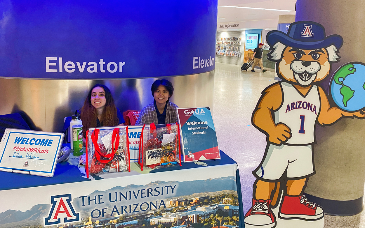 two female student Global Ambassadors at a table at the Wildcat Welcome Tucson Airport with a cutout of Wilbur Wildcat at right. 
