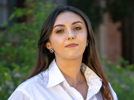 Photo of Montessa White, Visual Designer for Arizona International. They have brown hair and a white button up shirt. The background is blurred trees, shrubs and a red brick building. 