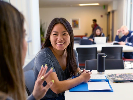 A female student smiles as another student talks to her.