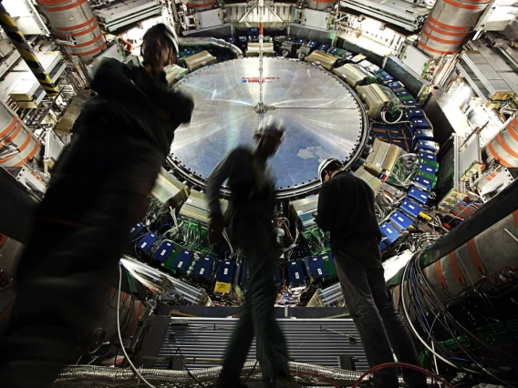 Three technicians in hard hats and work gear stand inside the vast, high-tech interior of the ATLAS detector at CERN’s Large Hadron Collider. The circular structure behind them is a massive, intricate assembly of wires, electronics, and machinery, with a large metallic disc at the center labeled “ATLAS CERN LHC.” The image conveys the scale and complexity of particle physics research and engineering. The lighting highlights the equipment, while the technicians appear slightly blurred, emphasizing motion and