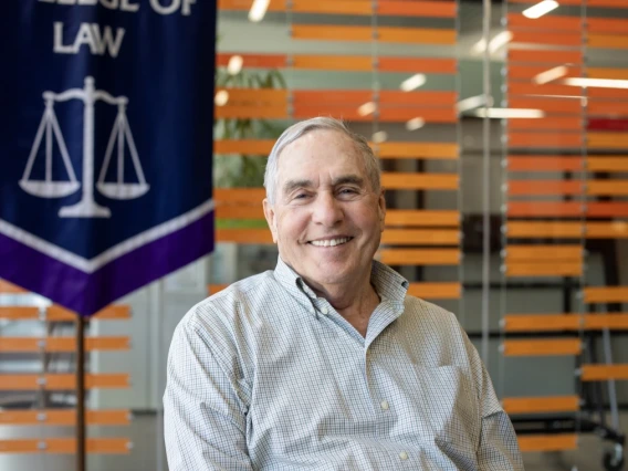 An older man with gray hair and a friendly smile sits in front of a modern backdrop of horizontal orange slats. He wears a light checkered button-down shirt and is positioned beside a banner displaying scales of justice and the words “College of Law,” suggesting a connection to a law school. The setting appears to be an academic or institutional space.