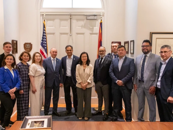 A group of fourteen individuals, including men and women, pose together in a formal setting in front of a pair of white double doors flanked by the U.S. and Arizona state flags. Most individuals are dressed in business attire, suggesting a diplomatic or professional delegation. In the foreground, a framed artwork rests on a wooden table, indicating a ceremonial exchange or gift presentation. The room is well-lit and adorned with framed certificates and plaques, reinforcing the official and celebratory natur