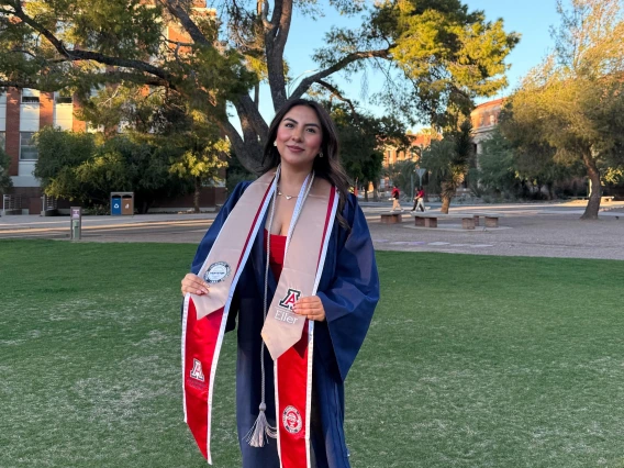 A young woman wearing a navy blue graduation gown and several stoles stands on a grassy area of the University of Arizona campus during golden hour. She smiles confidently, holding the ends of her stoles, which feature the University of Arizona, Eller College of Management, and other honors logos. She wears a red dress and white high-heeled sandals. Behind her, large trees and red-brick campus buildings are bathed in warm sunlight, creating a celebratory and serene atmosphere.