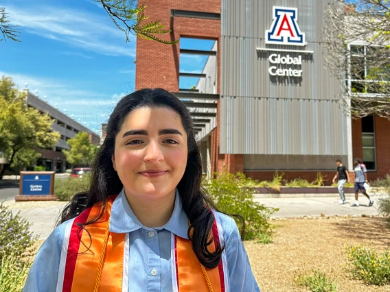 A young woman wearing a light blue shirt and an orange and red graduation stole smiles in front of the University of Arizona Global Center. The building behind her prominently features the U of A logo and the words “Global Center” on a modern metal and brick facade. Trees, desert landscaping, and a few students walking in the background reflect a sunny day on campus, highlighting a celebratory graduation moment.