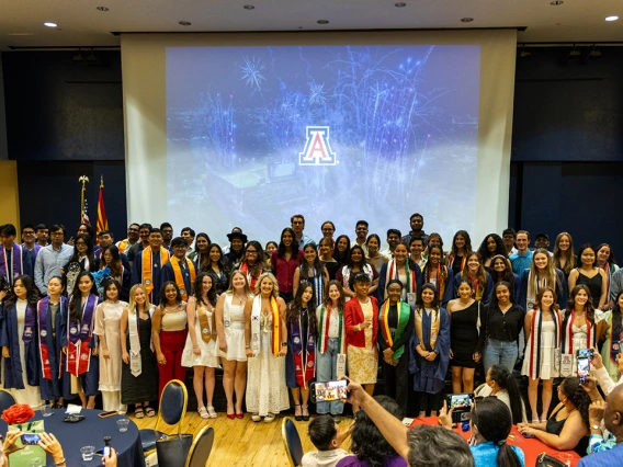 A large group of graduating students pose together on stage at the University of Arizona’s Global Graduation celebration. The graduates wear a mix of academic regalia, including caps, gowns, and colorful stoles representing international programs and cultural affiliations. Behind them, a large projection screen displays fireworks and the U of A logo. The event takes place in a decorated banquet hall with audience members seated at round tables, capturing the moment on their phones. The atmosphere is festive