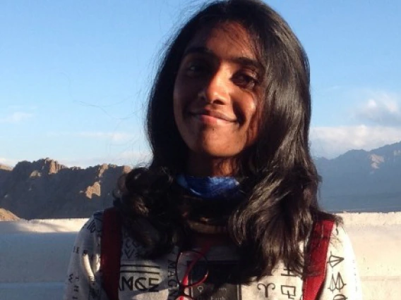 A young woman with long, dark hair smiles gently at the camera while standing outdoors in front of a mountainous landscape under a clear blue sky. She wears a white top with black printed symbols and a blue neck gaiter, with red backpack straps visible over her shoulders. The warm sunlight casts a soft glow on her face, suggesting a peaceful and scenic moment during travel or study abroad.
