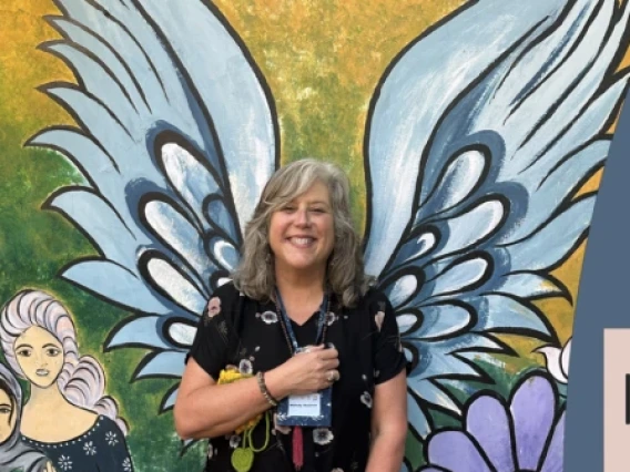 A smiling woman stands in front of a colorful mural featuring large angel wings and floral designs, creating the illusion that she has wings. She holds a pamphlet and wears a University of Arizona lanyard. To the right, a graphic overlay reads: “International education and its invaluable relevance” with a subheading, “A powerful message from the University of Arizona’s Melody Buckner and her Fulbright experience in India.” The design is branded with the U of A Research, Innovation & Impact logo, promoting g