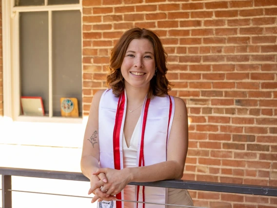 A young woman wearing a white dress and red-and-white graduation stole stands smiling and leaning on a railing in front of a red brick wall. Her stoles feature the University of Arizona and College of Humanities logos, as well as honor cords. Sunlight filters in from the left, casting a warm glow over the scene and highlighting her achievement and cheerful expression. A tattoo is visible on her upper arm, and framed mementos can be seen on the windowsill behind her.