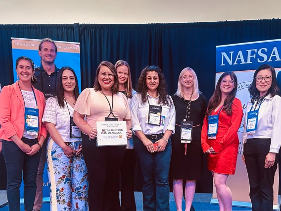 A group of international education professionals stands together on stage at the NAFSA conference, smiling for the camera. They are posed in front of blue and orange NAFSA banners, with one person holding a sign thanking the University of Arizona for serving as a table host. The attendees wear conference badges, and the atmosphere reflects a celebratory and collaborative event.