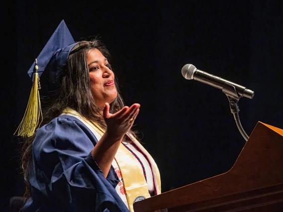 Ashwini R. Jannu, MSIS ’25, delivers the graduate student speech at the Spring 2025 College of Information Science Convocation. Wearing a cap and gown with a gold stole and honor cords, she gestures expressively while speaking into a microphone from behind a podium. Jannu was honored as the Outstanding Graduate Student award-winner.