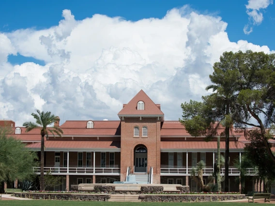 A front-facing view of Old Main, the historic red-brick building at the University of Arizona, framed by palm trees and desert greenery. A wide staircase leads to the main entrance under a prominent gabled roof. Puffy white clouds and a bright blue sky form a dramatic backdrop, highlighting the architectural charm and heritage of the university’s oldest building.