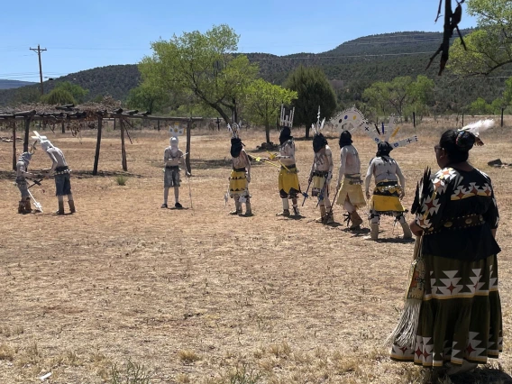 A group of Indigenous dancers dressed in traditional attire perform an outdoor ceremonial dance on dry grassland surrounded by trees and mesas. Male dancers wear body paint, headpieces, and yellow garments, while female dancers in colorful skirts and sashes observe or participate. The scene is set in a rural landscape under a clear blue sky.