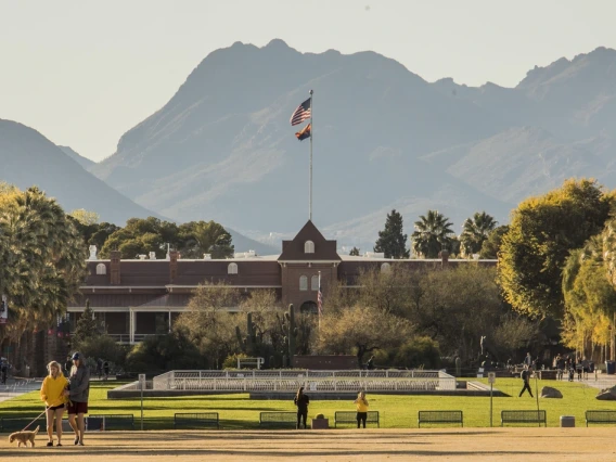 View of Old Main at the University of Arizona with the American and Arizona state flags flying atop the building. The scene is set against the backdrop of the Santa Catalina Mountains, with students walking and relaxing on the grass in the foreground, surrounded by palm trees and desert vegetation.