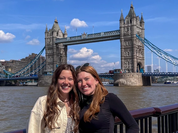 Two women smiling and standing close together in front of London’s Tower Bridge on a sunny day, with the River Thames and the iconic twin towers of the bridge in the background.