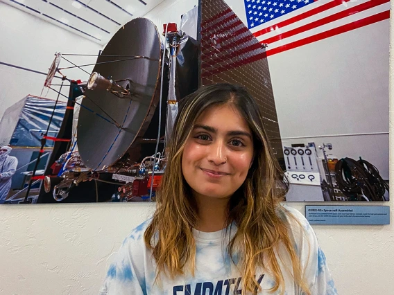Maanyaa Kapur, wearing a light blue tie-dye “EMPATHY” T-shirt, smiles while standing indoors in front of a large photograph of the OSIRIS-REx spacecraft assembly, which features a satellite dish and an American flag.