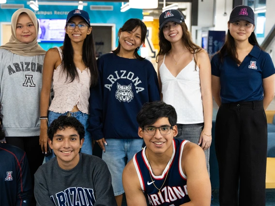 Eight students pose together indoors, all wearing University of Arizona apparel, including shirts, sweatshirts, and hats. Five students stand in the back row, and three kneel in the front, smiling at the camera. The background features a brightly lit lounge area with blue seating and a “Campus Recreation” sign.
