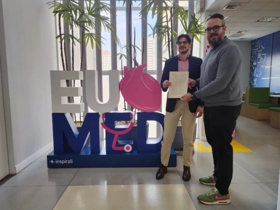 Two men stand indoors in front of a large “EU MED” sign, holding a signed document together and smiling. The sign features a pink anatomical heart and stethoscope design, with potted plants and tall windows in the background.