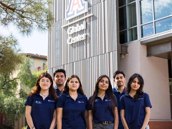 A group of seven University of Arizona Global Ambassadors stand together outside the Global Center, smiling at the camera. They are wearing matching navy polo shirts with the Arizona International logo, with the building’s large "A" logo and "Global Center" signage visible behind them.