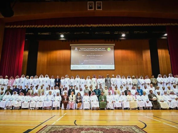 Members of the University of Arizona Gulf Cooperation Council (GCC-UA) alumni community pose for a large group photo on stage during the 11th Annual GCC Alumni Reunion in Salalah, Oman. The audience includes alumni, officials, and guests seated and standing in several rows inside an auditorium decorated with red curtains and projection screens.