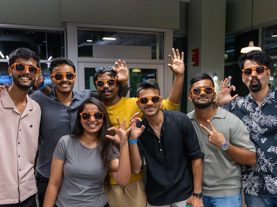 A group of seven students wearing matching orange sunglasses smile and pose together at an indoor event. They make hand gestures, including the University of Arizona "Wildcat" hand sign, while enjoying the social atmosphere.