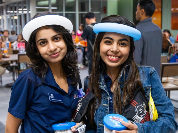 Two University of Arizona students wearing balloon hats and navy Arizona International polo shirts smile while holding ice cream and snacks at a student event inside the Global Center.