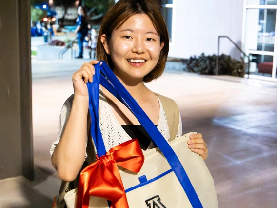 A student smiles while holding a University of Arizona Campus Recreation tote bag decorated with a large red bow during an evening event.