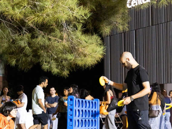 Students play a giant Connect Four game on the lawn outside the University of Arizona Global Center during a nighttime event, with other attendees mingling in the background.