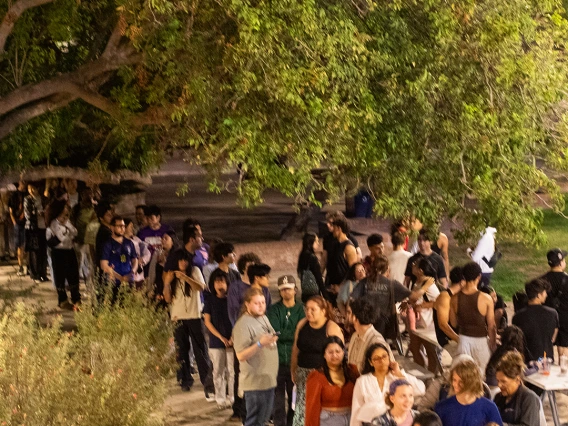 A large group of students lines up outdoors under the trees at night during a campus event. In the foreground, people gather around tables with red tablecloths, while the line of attendees stretches back into the distance. The scene is brightly lit, highlighting the energy and turnout of the event.