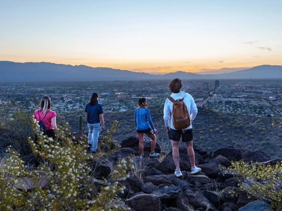 Four people stand on rocky terrain at sunset, overlooking the city of Tucson, Arizona. The view includes the sprawling cityscape below with distant mountains and a colorful sky.
