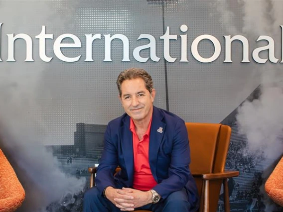 Hassan Hijazi, Senior Assistant Vice President for University of Arizona International, sits in a chair in the Arizona International office. He is wearing a navy blue suit with a red shirt, smiling at the camera. Behind him, the word “International” is displayed prominently on the wall.