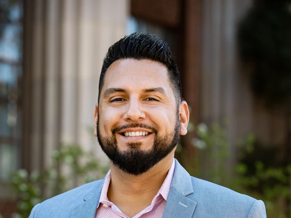 Portrait of Ricky Salazar smiling outdoors in front of a building with columns. He is wearing a light blue blazer and a pink collared shirt.