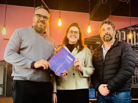 Three people stand together indoors under warm hanging lights, smiling at the camera. The person on the left holds a purple and blue booklet. The background features a chalkboard-style mural with text and illustrations, giving the space a casual, creative atmosphere.