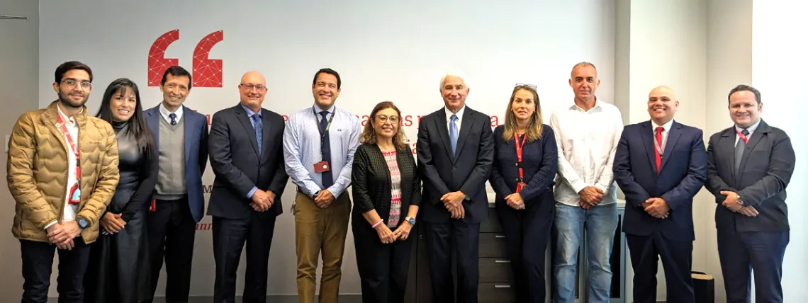 Leadership from UPC and UArizona are standing in a row, smiling at the camera. They are dressed in business attire, ranging from suits and ties to business casual clothing. The background features a white wall with red text and symbols.