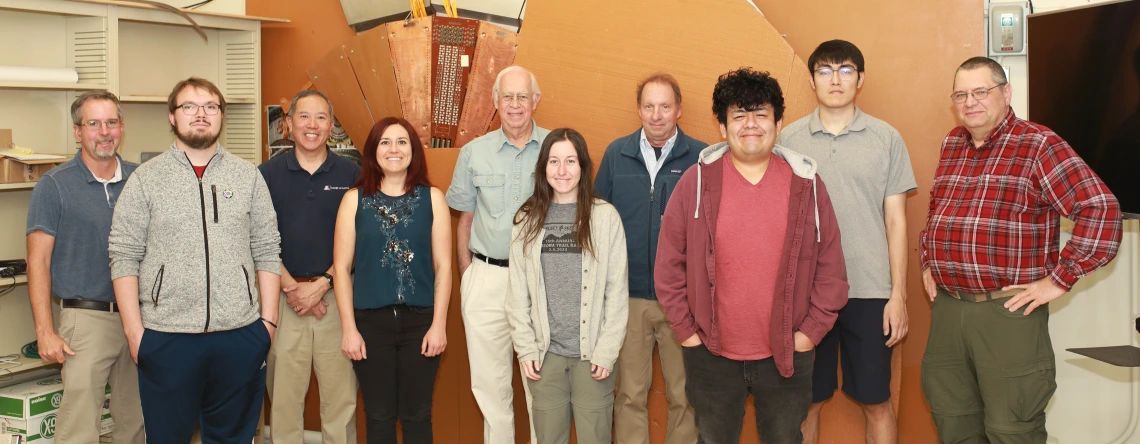 A group of eleven individuals—comprising a mix of students, faculty, and researchers—stand side by side smiling for a photo in a lab or workspace. Behind them is a large, intricate copper-colored component, likely a particle detector or scientific instrument. The group appears to be part of a research team, possibly involved with the ATLAS experiment or related particle physics project. They are dressed in casual and business-casual attire, and the environment reflects an academic or engineering setting.