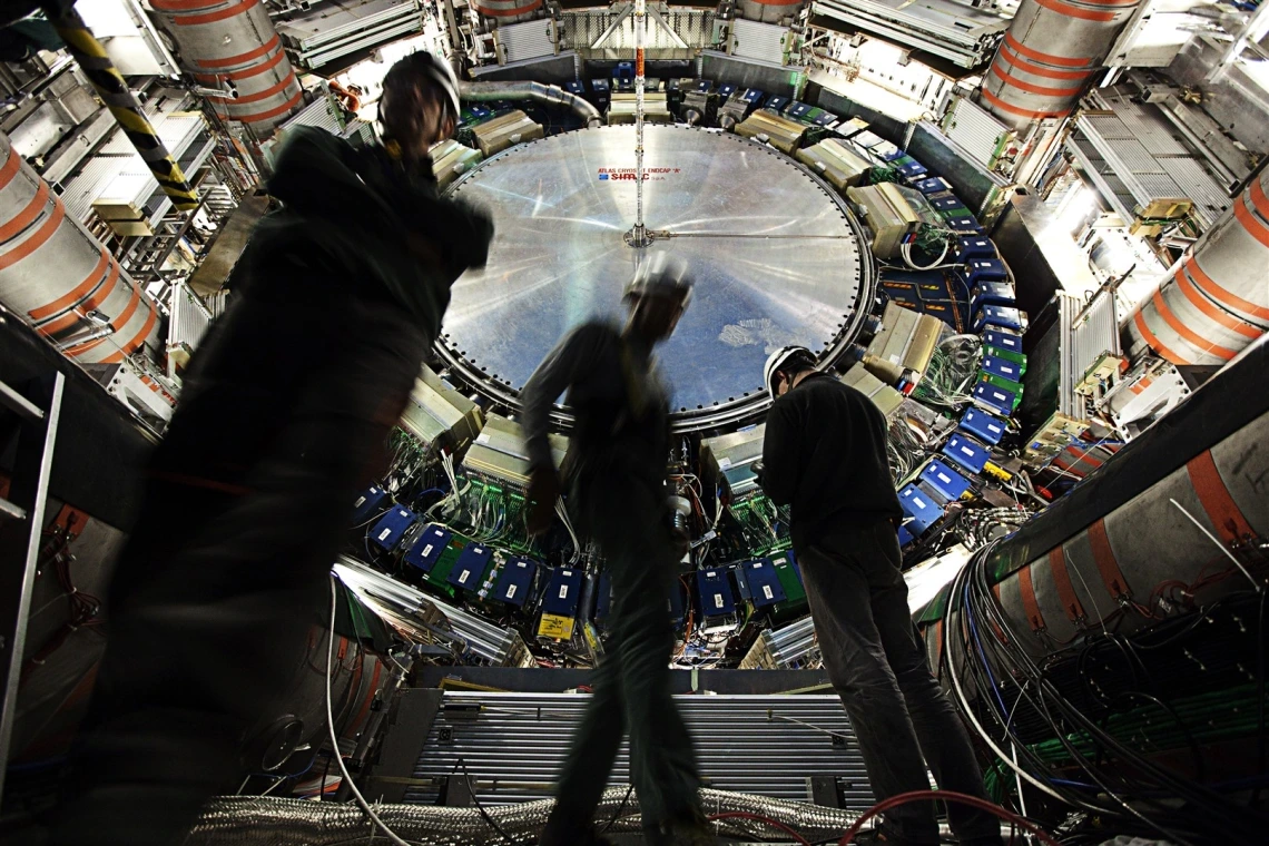 Three technicians in hard hats and work gear stand inside the vast, high-tech interior of the ATLAS detector at CERN’s Large Hadron Collider. The circular structure behind them is a massive, intricate assembly of wires, electronics, and machinery, with a large metallic disc at the center labeled “ATLAS CERN LHC.” The image conveys the scale and complexity of particle physics research and engineering. The lighting highlights the equipment, while the technicians appear slightly blurred, emphasizing motion and
