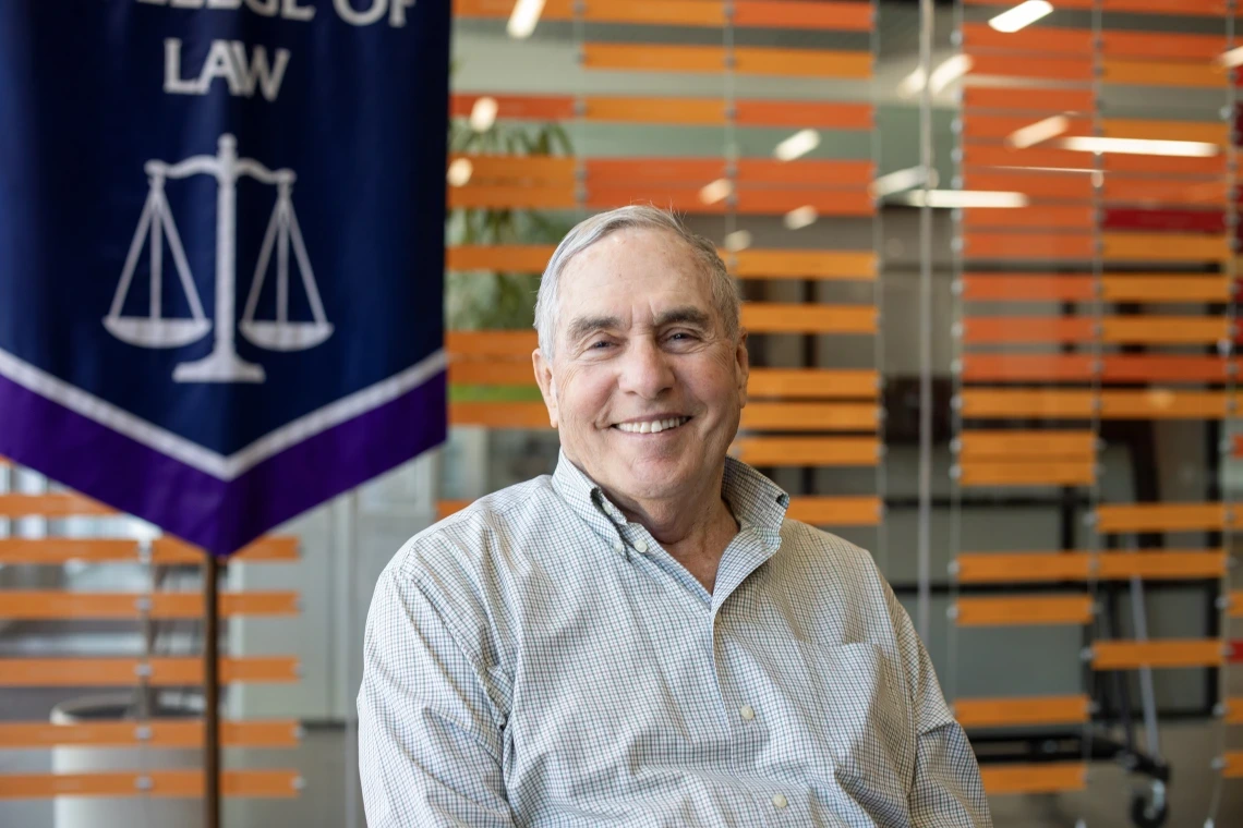 An older man with gray hair and a friendly smile sits in front of a modern backdrop of horizontal orange slats. He wears a light checkered button-down shirt and is positioned beside a banner displaying scales of justice and the words “College of Law,” suggesting a connection to a law school. The setting appears to be an academic or institutional space.