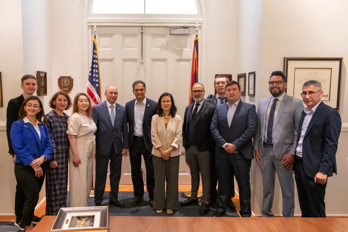 A group of fourteen individuals, including men and women, pose together in a formal setting in front of a pair of white double doors flanked by the U.S. and Arizona state flags. Most individuals are dressed in business attire, suggesting a diplomatic or professional delegation. In the foreground, a framed artwork rests on a wooden table, indicating a ceremonial exchange or gift presentation. The room is well-lit and adorned with framed certificates and plaques, reinforcing the official and celebratory natur