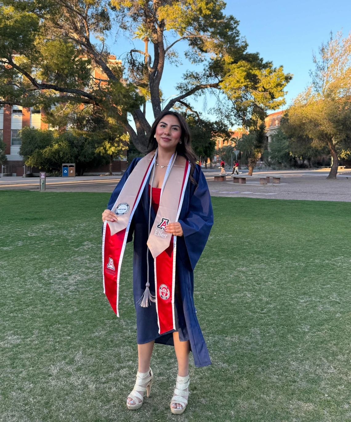 A young woman wearing a navy blue graduation gown and several stoles stands on a grassy area of the University of Arizona campus during golden hour. She smiles confidently, holding the ends of her stoles, which feature the University of Arizona, Eller College of Management, and other honors logos. She wears a red dress and white high-heeled sandals. Behind her, large trees and red-brick campus buildings are bathed in warm sunlight, creating a celebratory and serene atmosphere.