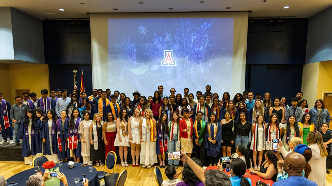 A large group of graduating students pose together on stage at the University of Arizona’s Global Graduation celebration. The graduates wear a mix of academic regalia, including caps, gowns, and colorful stoles representing international programs and cultural affiliations. Behind them, a large projection screen displays fireworks and the U of A logo. The event takes place in a decorated banquet hall with audience members seated at round tables, capturing the moment on their phones. The atmosphere is festive