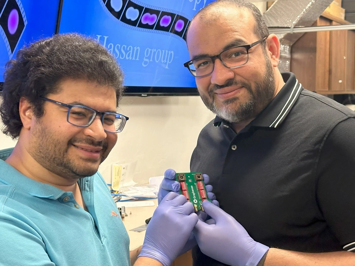 Two researchers smile while holding a small green circuit board labeled “Graphene” in a laboratory setting. Both are wearing glasses and blue gloves, with one in a teal polo shirt and the other in a black collared shirt. A monitor in the background displays scientific imagery and the words “Hassan group.” The setting suggests an academic research lab focused on advanced electronics or materials science.