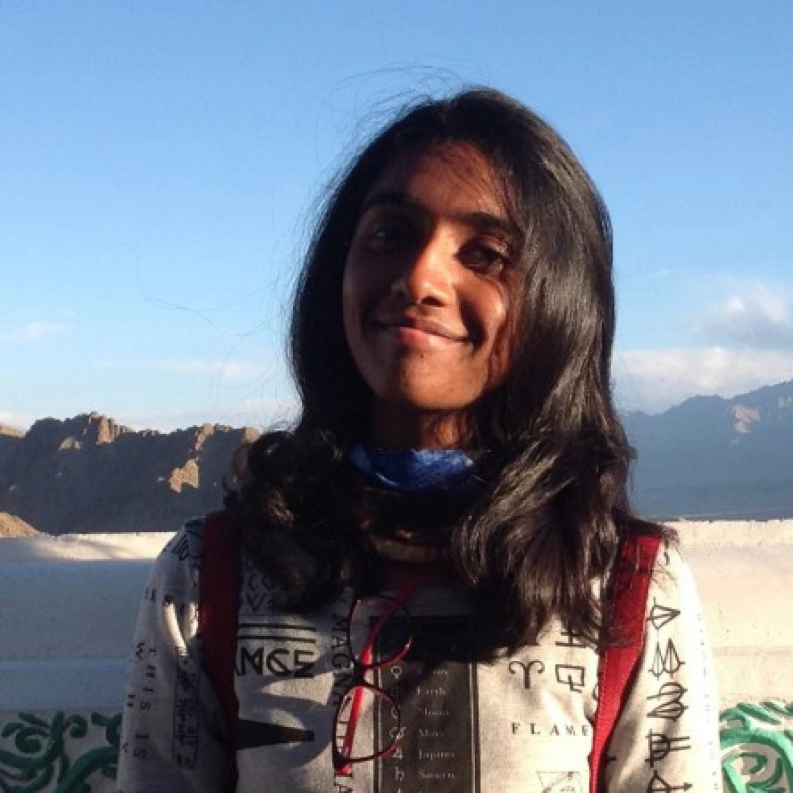 A young woman with long, dark hair smiles gently at the camera while standing outdoors in front of a mountainous landscape under a clear blue sky. She wears a white top with black printed symbols and a blue neck gaiter, with red backpack straps visible over her shoulders. The warm sunlight casts a soft glow on her face, suggesting a peaceful and scenic moment during travel or study abroad.