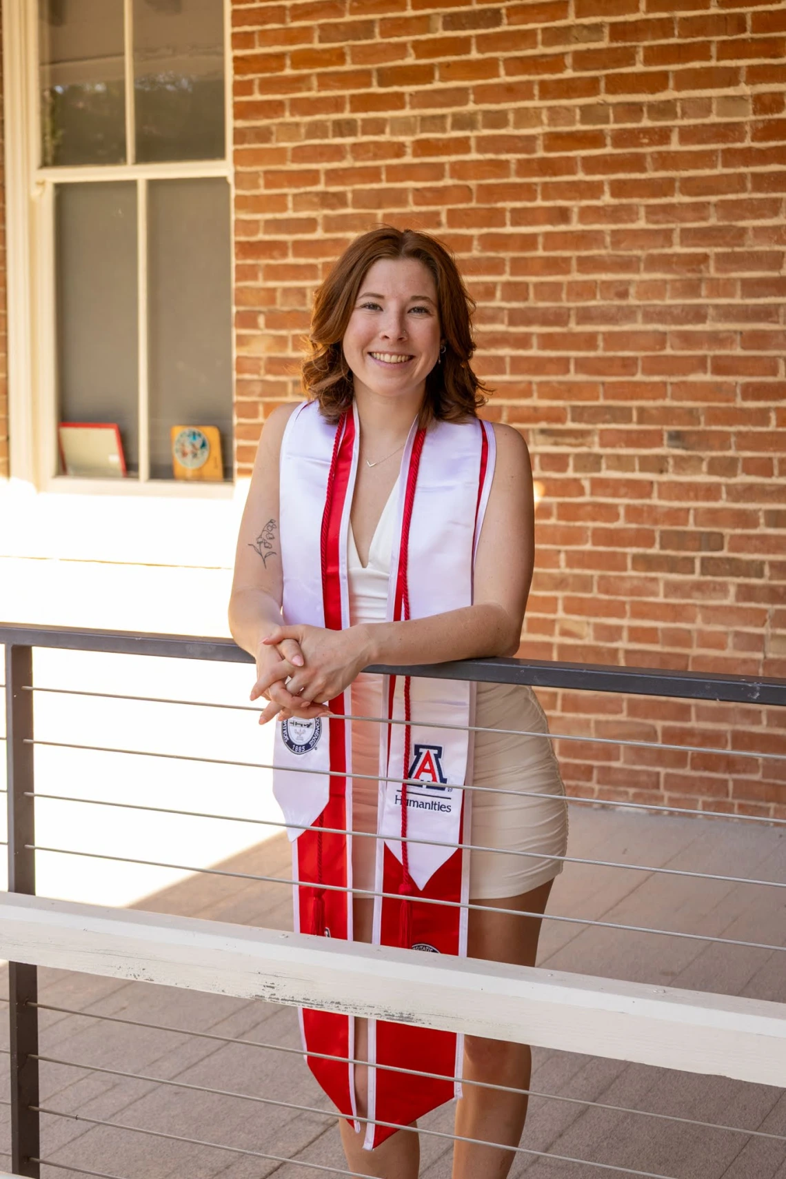 A young woman wearing a white dress and red-and-white graduation stole stands smiling and leaning on a railing in front of a red brick wall. Her stoles feature the University of Arizona and College of Humanities logos, as well as honor cords. Sunlight filters in from the left, casting a warm glow over the scene and highlighting her achievement and cheerful expression. A tattoo is visible on her upper arm, and framed mementos can be seen on the windowsill behind her.