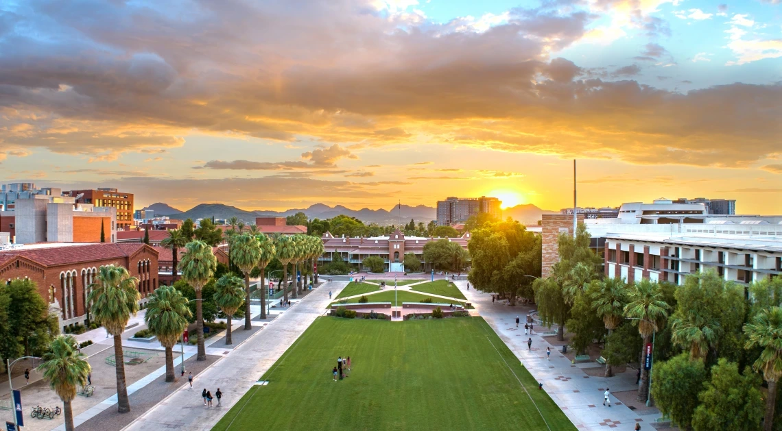 A wide view of the University of Arizona campus at golden hour, looking down the central grassy Mall lined with palm trees. Students are walking and gathering along the paths. Historic red-brick and modern academic buildings flank the green lawn, with a backdrop of mountain silhouettes and a dramatic sunset sky filled with warm orange and purple hues. The setting sun casts a golden glow across the scene, highlighting the campus's vibrant and welcoming atmosphere.