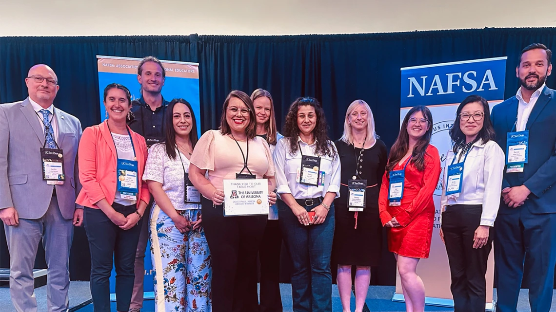 A group of international education professionals stands together on stage at the NAFSA conference, smiling for the camera. They are posed in front of blue and orange NAFSA banners, with one person holding a sign thanking the University of Arizona for serving as a table host. The attendees wear conference badges, and the atmosphere reflects a celebratory and collaborative event.