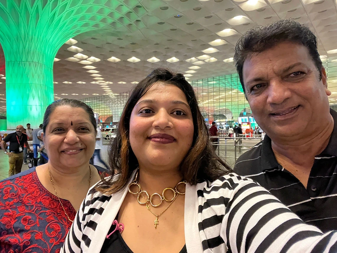 Ashwini R. Jannu takes a smiling selfie with her parents at an airport, standing in front of a brightly lit green column beneath a modern, geometric ceiling.