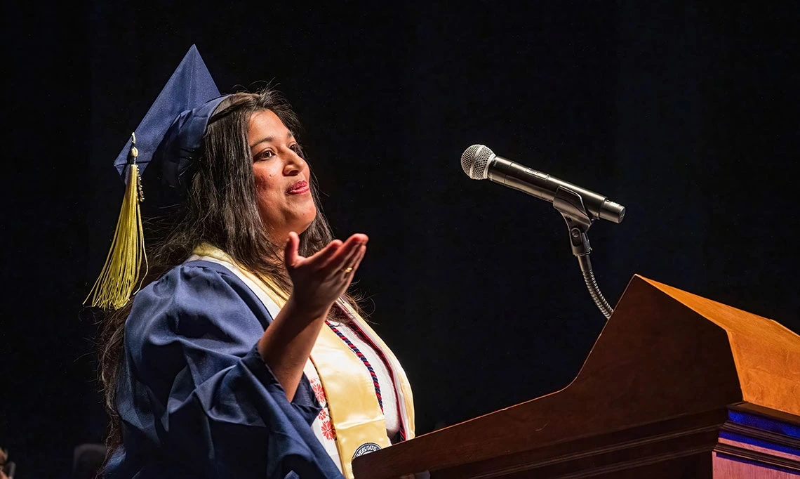 Ashwini R. Jannu, MSIS ’25, delivers the graduate student speech at the Spring 2025 College of Information Science Convocation. Wearing a cap and gown with a gold stole and honor cords, she gestures expressively while speaking into a microphone from behind a podium. Jannu was honored as the Outstanding Graduate Student award-winner.