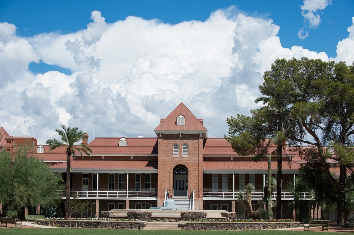 A front-facing view of Old Main, the historic red-brick building at the University of Arizona, framed by palm trees and desert greenery. A wide staircase leads to the main entrance under a prominent gabled roof. Puffy white clouds and a bright blue sky form a dramatic backdrop, highlighting the architectural charm and heritage of the university’s oldest building.