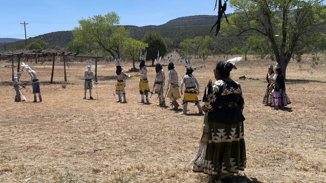 A group of Indigenous dancers dressed in traditional attire perform an outdoor ceremonial dance on dry grassland surrounded by trees and mesas. Male dancers wear body paint, headpieces, and yellow garments, while female dancers in colorful skirts and sashes observe or participate. The scene is set in a rural landscape under a clear blue sky.